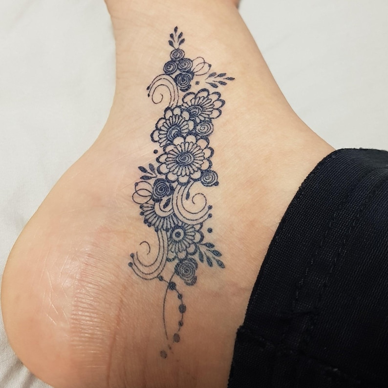 May include: A dark blue floral tattoo design on the side of a foot. The tattoo features a series of flowers, swirls, and dots. The design is intricate and detailed, with a focus on the natural curves of the foot.