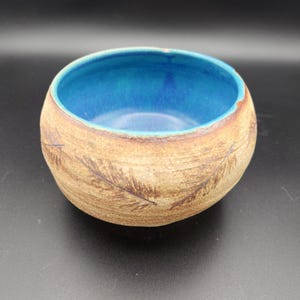 May include: A small, round ceramic bowl with a textured, light brown exterior featuring dark brown feather imprints. The interior of the bowl is glazed in a vibrant, glossy blue, creating a distinct contrast with the earthy, matte outer surface.