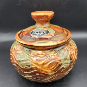 May include: A round ceramic lidded jar with a glossy brown and green marbled glaze. The lid features a central knob and a black oval sticker that reads Dryden Potter's Wheel ORIGINAL. The body has a textured, wavy, and ridged surface finish.