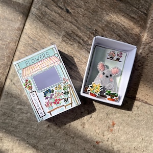 以下が含まれることがあります： 2つの小さな白い箱。1つの箱には、上部に「FLOWERS」、側面に「BLOOMS」という文字が入った花屋の絵が描かれています。もう1つの箱には、ピンクの耳を持つ灰色のマウスとフラワーアレンジメントが入っています。
