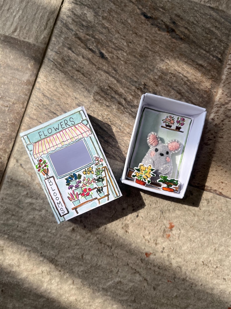 以下が含まれることがあります： 2つの小さな白い箱。1つの箱には、上部に「FLOWERS」、側面に「BLOOMS」という文字が入った花屋の絵が描かれています。もう1つの箱には、ピンクの耳を持つ灰色のマウスとフラワーアレンジメントが入っています。
