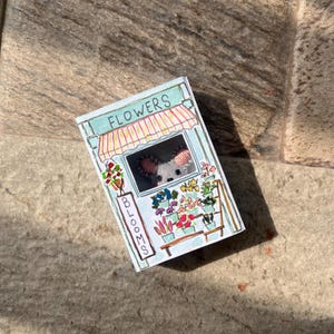 以下が含まれることがあります： 手描きの小さな花屋のイラスト。上部に「FLOWERS」、側面に「BLOOMS」の文字。お店にはストライプのオーニングがあり、色とりどりの鉢植えの花でいっぱいです。コアラがお店の窓から覗いています。