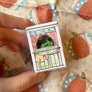 以下が含まれることがあります： 手描きの小さな紙製ジオラマで、バルコニーのシーンを描いています。バルコニーは緑色に塗られており、赤いカーテンと黄色のショートパンツが物干しロープに吊るされています。背景は、オレンジ色のカボチャが描かれたベージュ色の生地です。