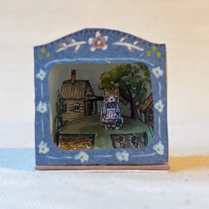 Pode incluir: Um diorama em miniatura com um personagem de gato em uma cena de jardim. A moldura azul é decorada com motivos florais. A cena inclui uma casa, árvores e canteiros. O gato está usando um avental.
