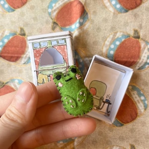 以下が含まれることがあります： 手作りの緑色のフェルトカエルのピンバッジ。黒いビーズのアクセント付き。小さなイラスト入りの箱の近くにあります。箱にはキッチンのシーンの絵が描かれています。気まぐれなアクセサリーです。