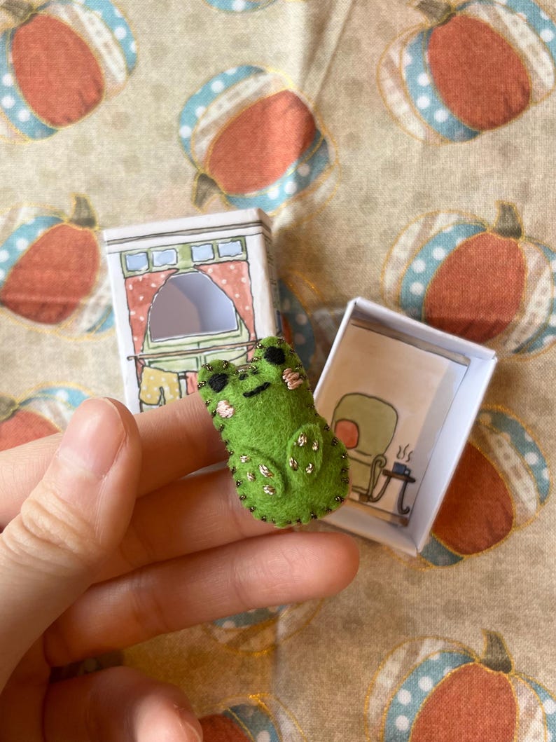 以下が含まれることがあります： 手作りの緑色のフェルトカエルのピンバッジ。黒いビーズのアクセント付き。小さなイラスト入りの箱の近くにあります。箱にはキッチンのシーンの絵が描かれています。気まぐれなアクセサリーです。
