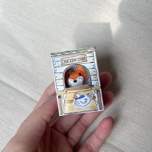 以下が含まれることがあります： 手描きのミニチュア鶏小屋で、キツネが覗いています。 小屋には「CHICKEN COOP」の文字と、描かれた鶏が描かれています。 キツネはオレンジ色の毛皮と白い顔をしています。 小屋は手で持たれています。