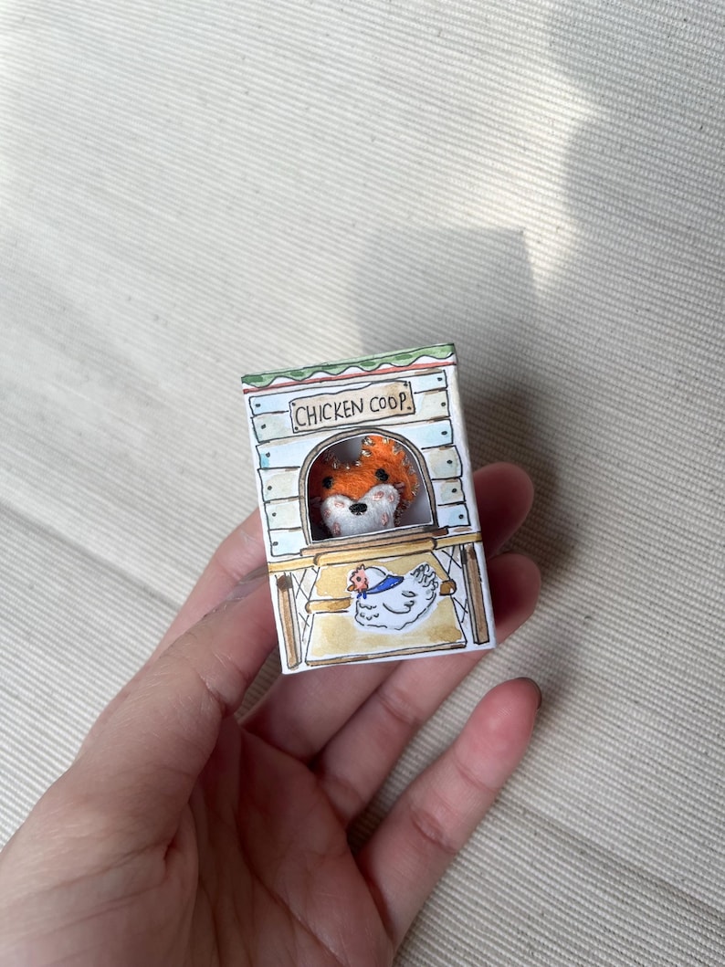 以下が含まれることがあります： 手描きのミニチュア鶏小屋で、キツネが覗いています。 小屋には「CHICKEN COOP」の文字と、描かれた鶏が描かれています。 キツネはオレンジ色の毛皮と白い顔をしています。 小屋は手で持たれています。