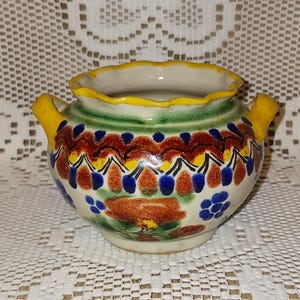 May include: A small, hand-painted ceramic pot with a scalloped yellow rim and two yellow handles. The pot features a colorful design with blue, red, and brown floral patterns. The pot is sitting on a white lace tablecloth.