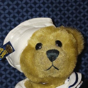 May include: A plush teddy bear wearing a white sailor hat and a navy blue and white striped collar. The bear has a tan fur coat, black eyes, and a black nose. A small tag on the hat reads "20th Century".