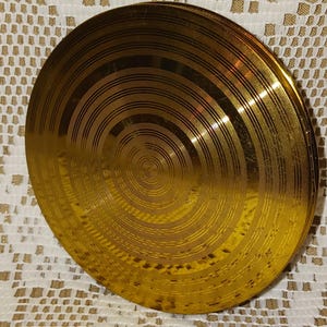 May include: A gold-colored, circular object with concentric circular grooves. The object has a shiny, reflective surface and appears to be made of metal. The design is reminiscent of a record or a decorative plate.