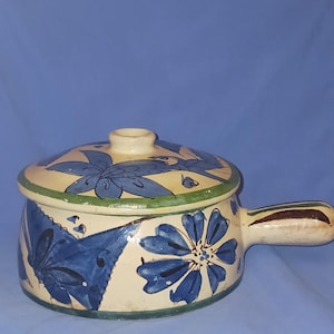 Puede incluir: Cazuela de cerámica color crema con tapa, con diseños florales azules y un asa. El plato tiene un borde verde y un asa marrón. Los patrones florales están pintados a mano, agregando un toque único a este artículo de cocina.