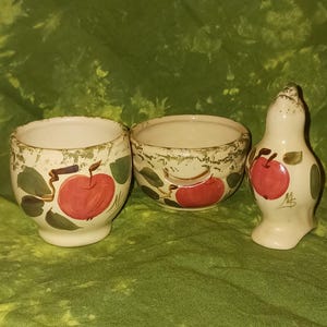May include: A set of three ceramic pieces: two small bowls and a salt shaker. Each piece is off-white with a hand-painted design of a red apple, green leaves, and brown stems. The set is on a green background.