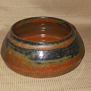 May include: A handmade ceramic bowl with a wide, rounded shape. The bowl features a rustic design with bands of color, including rust, dark blue, and green. The interior is a warm, reddish-brown color.