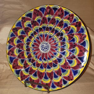May include: A round ceramic plate featuring a vibrant, hand-painted mandala pattern. The design consists of concentric layers of petal-like shapes in deep burgundy, royal blue, and bright yellow, centered around a small, circular grid motif.