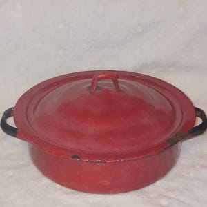 May include: A vintage red enamelware pot with a matching lid. The pot has a rounded shape with two black handles and a small handle on the lid. The enamel is chipped in places, giving it a rustic appearance. A classic kitchenware item.