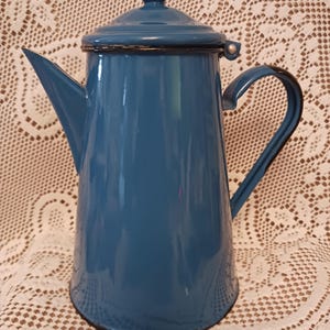 Puede incluir: Una cafetera vintage de esmalte azul con tapa y asa. La cafetera tiene un diseño clásico con un pico para verter. El asa y el borde son negros. La cafetera probablemente se utiliza para preparar café u otras bebidas calientes.