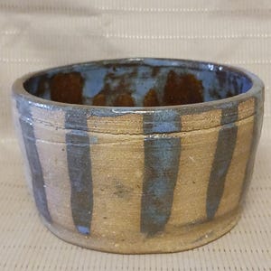 May include: A handmade ceramic bowl with a beige exterior and a blue interior. Vertical blue stripes adorn the outside. The bowl's rim is a darker shade of blue, and the interior has brown accents. This rustic bowl is suitable for serving food.