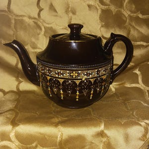 May include: A dark brown ceramic teapot with a lid and handle. The teapot features a decorative band with gold, white, and black patterns. The spout and handle are curved. The teapot is set against a gold-colored background.