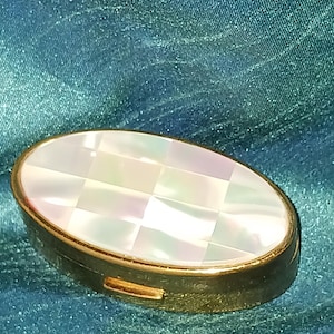 May include: An oval-shaped metal pill box with a gold-toned finish and a hinged lid. The top of the lid features an inlaid iridescent mother-of-pearl panel arranged in a grid pattern of small, reflective, light-catching squares.