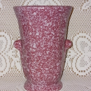 Puede incluir: Un jarrón moteado de color granate con una abertura ancha y dos asas pequeñas. El jarrón tiene una base redondeada y una superficie texturizada. El fondo es un mantel crema con dibujos. Este jarrón decorativo es adecuado para arreglos florales.