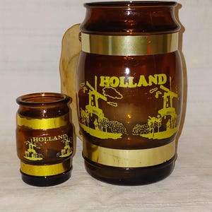 Peut inclure: Une chope à bière en verre brun et un verre à shot assorti, tous deux dotés de bandes métalliques dorées et de motifs sérigraphiés jaunes représentant des moulins à vent avec le texte HOLLAND. La chope possède une anse en bois clair.