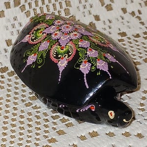 Könnte beinhalten: Eine schwarze Schildkrötenfigur mit glänzender Oberfläche, die auf ihrem Panzer ein detailliertes, handgemaltes Mandalamuster in Lavendel-, Rosa- und Grüntönen aufweist. Die Schildkröte hat einen kleinen Kopf mit bemalten Augen und ein gemustertes Gehäuse.