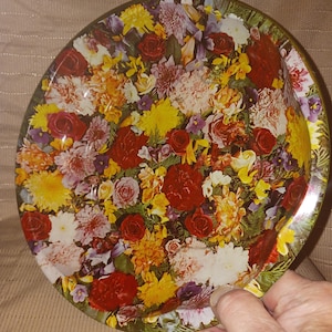 Puede incluir: Una bandeja redonda de metal con un patrón floral denso y colorido. El diseño incluye una variedad de flores en tonos rojos, amarillos, naranjas, blancos y morados, dispuestas en una composición apretada y superpuesta en toda la superficie.