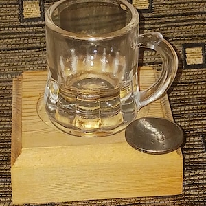 Puede incluir: Una pequeña jarra de cristal transparente con asa, apoyada sobre una base de madera clara. Una pequeña tapa redonda de metal está al lado de la jarra. La jarra tiene un diseño acanalado.