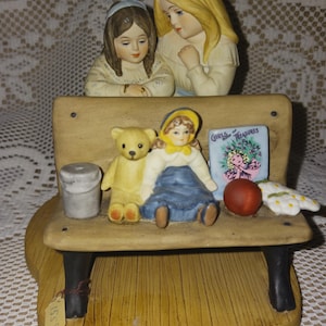 Könnte beinhalten: Keramik-Bankfigur mit zwei Mädchen, einem Teddybär und einer Puppe. Die Mädchen sitzen auf der Bank, mit der Puppe und dem Bären neben sich. Ein kleines Buch mit den Worten "Girls Treasures" befindet sich ebenfalls auf der Bank.