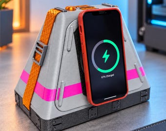 May include: A grey, triangular wireless charging dock with a bright orange strap and a horizontal hot pink stripe. A smartphone with a red case is attached to the front, displaying a green circular charging icon and the text 67% Charged.