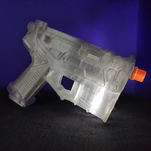 May include: A transparent, plastic toy gun with an orange barrel. The toy gun is a detailed replica of a firearm, featuring various molded components and textures. The gun is set against a dark surface and a blue background.