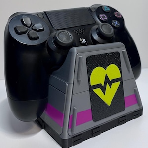 May include: A black PlayStation 4 controller on a grey charging stand. The stand has a yellow heart design with a lightning bolt, and a purple stripe. The controller has black buttons and joysticks.