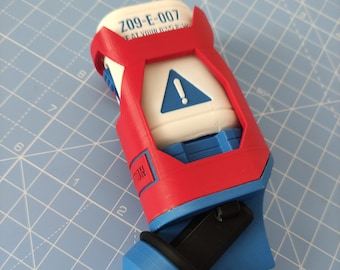 Può includere: Un dispositivo stampato in 3D rosso e blu con un contenitore bianco etichettato "Z09-E-007 FAT YOUR OWN RISK" e un cartello di avvertimento blu con un punto esclamativo. Il dispositivo ha un'impugnatura nera e un ugello blu.