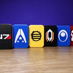 May include: A row of six rectangular metal tins featuring various graphic designs. The first is black with the white and red N7 logo, followed by a blue tin with a white star emblem, a yellow tin with a black sun icon, a black tin with an orange and white geometric shape, a blue tin with a white circle, and a red tin with a white skull graphic.