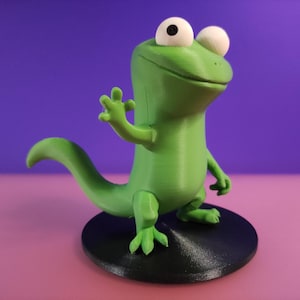 Könnte beinhalten: Eine grüne Eidechsenfigur mit großen weißen Augen und einem freundlichen Gesichtsausdruck. Die Eidechse steht auf einem schwarzen, kreisförmigen Sockel vor einem lila und rosa Hintergrund. Die Eidechse winkt mit einer Hand.