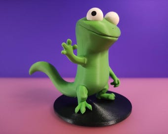 Pode incluir: Uma estatueta de lagarto verde com grandes olhos brancos e uma expressão amigável. O lagarto está em pé sobre uma base circular preta contra um fundo roxo e rosa. O lagarto está acenando com uma mão.