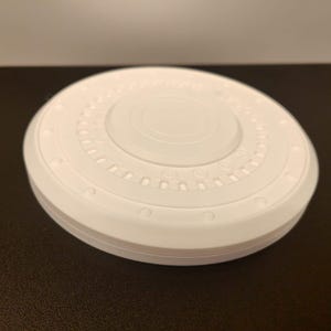 May include: A white, circular object with a layered design. The top surface features concentric circles and a series of small, evenly spaced indentations. The object is sitting on a dark surface.