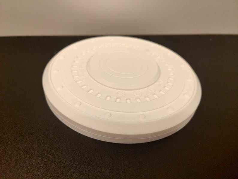 May include: A white, circular object with a layered design. The top surface features concentric circles and a series of small, evenly spaced indentations. The object is sitting on a dark surface.