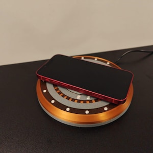 May include: A red smartphone rests on a wireless charging pad. The pad has a circular design with copper and gray accents, and small white lights. A black cable extends from the pad.