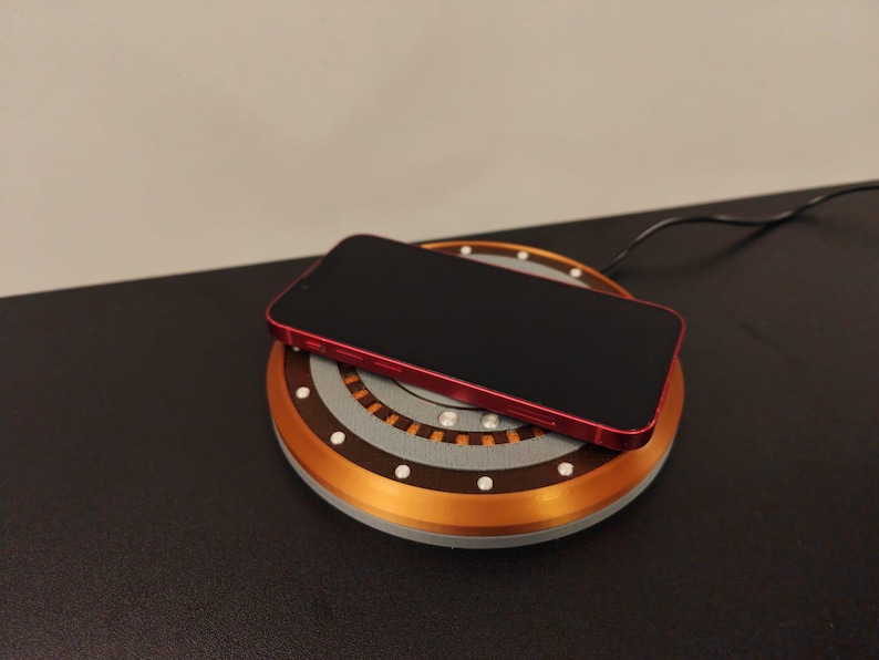 May include: A red smartphone rests on a wireless charging pad. The pad has a circular design with copper and gray accents, and small white lights. A black cable extends from the pad.