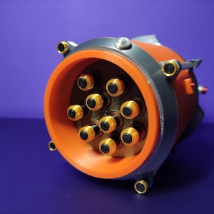 Könnte beinhalten: Nahaufnahme eines orangefarbenen und grauen Industrie-Steckverbinders mit mehreren goldfarbenen Stiften. Der kreisförmige Stecker hat eine Reihe von schwarzen und goldenen Kontaktpunkten. Der Hintergrund ist einfarbig lila.