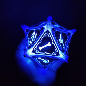 May include: A blue glowing 3D printed D20 dice with a white outline and black numbers. The dice is being held in a hand.