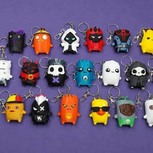 May include: A collection of seventeen small, colorful character keychains featuring various designs including a yellow banana, a red devil, a white ghost, a skull figure, a blue creature, a yellow duck with sunglasses, and other stylized figures.
