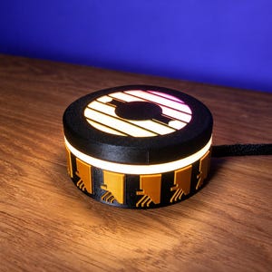 May include: A circular black device with a glowing top surface featuring a central black circle and horizontal light bars. The side of the device is black with a series of raised yellow rectangular accents, each with a small downward-pointing detail.