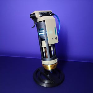 Puede incluir: Un dispositivo beige y negro con un gatillo, un tubo azul y un cuerpo cilíndrico. El dispositivo está montado sobre una base circular negra. El texto "Teddy Makers Flow Pen" es visible.