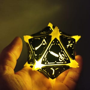 May include: A black and white illuminated 20-sided die with a geometric design. The die is held in a hand and is glowing yellow.