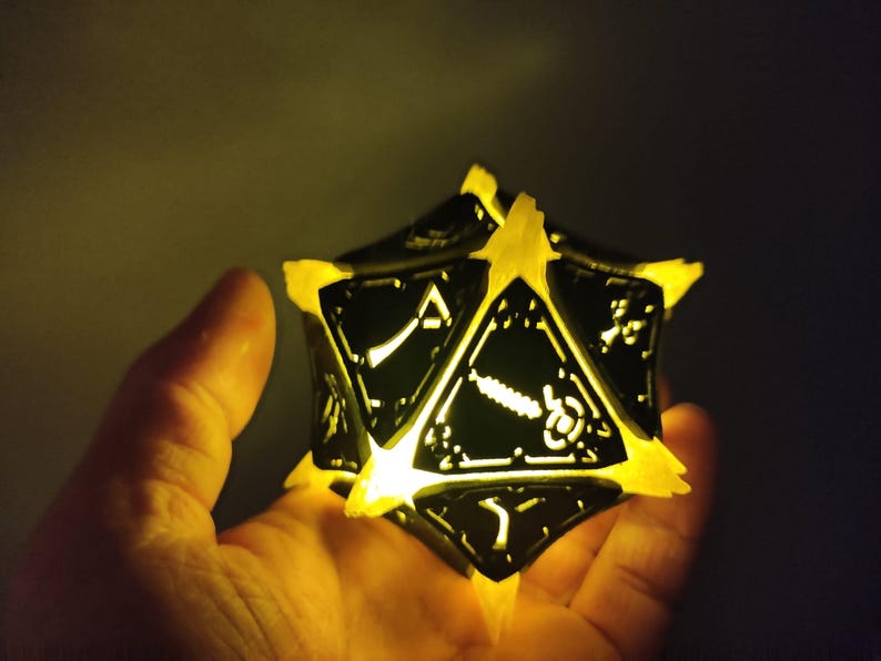 May include: A black and white illuminated 20-sided die with a geometric design. The die is held in a hand and is glowing yellow.