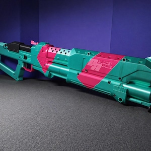 Puede incluir: Una pistola de juguete de color turquesa y rosa con el texto "BUDGET ARMS" en el lateral. La pistola de juguete tiene una punta naranja y está sobre un fondo morado y gris.