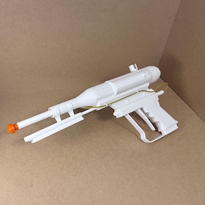 Może przedstawiać: Biała zabawka blaster z pomarańczową końcówką, z długą lufą, lunetą i uchwytem. Broń rekwizytowa ma futurystyczny design i jest wykonana z tworzywa sztucznego.